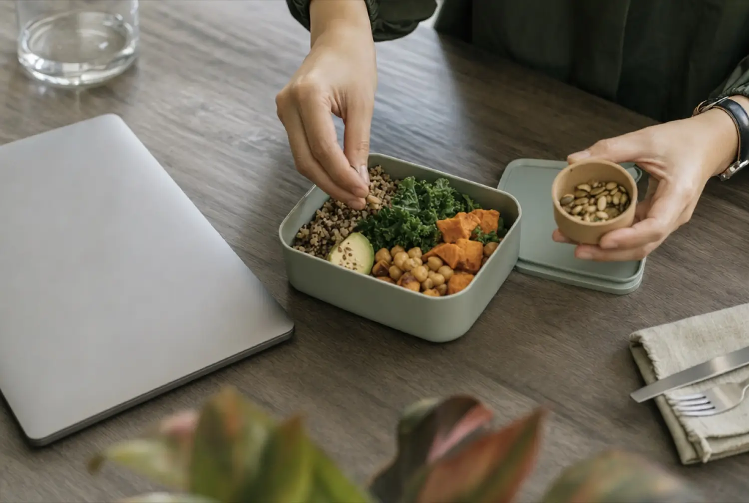 Hands assembling a lunch of grains and greens in a container on a meeting-room table beside a laptop